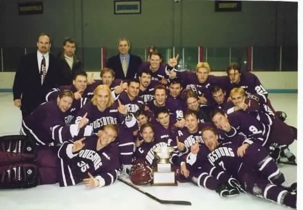 The Augsburg team in purple jerseys gathered on the ice around the championship trophy with the coaching staff standing behind